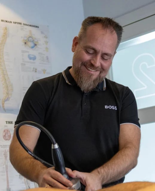 Man with beard wearing black BOSS polo shirt holding handheld device, with anatomical charts visible in background